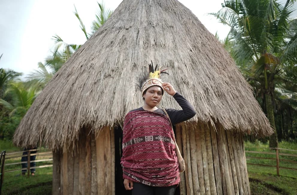 A person in patterned clothing and a headdress standing in front of a thatched-roof building